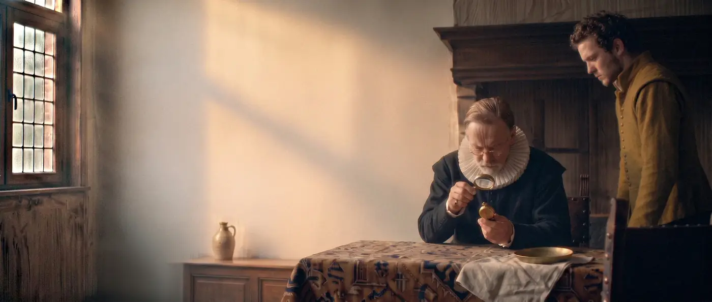 A merchant's room in Antwerp in 1610: a buyer studies a pocket watch through a magnifying glass while the seller waits beside a carpet-covered table.